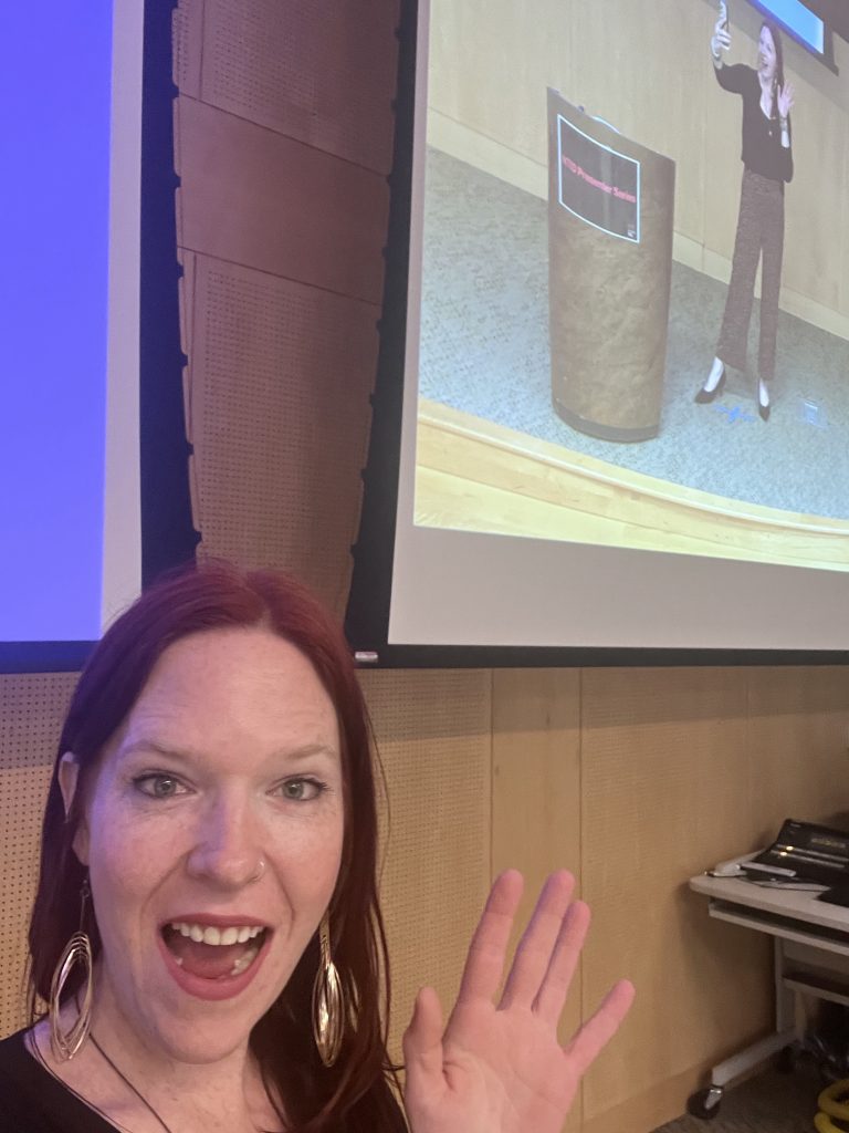 Rach smiling and waving at a speaking event, with her presentation on the screen behind her